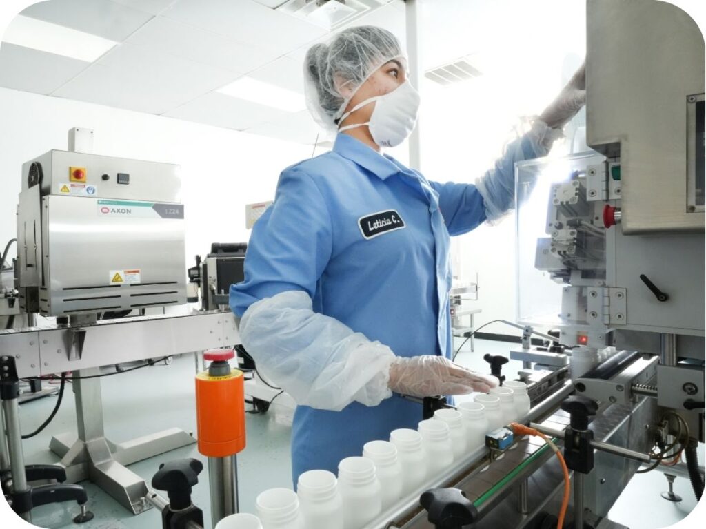 A cleanroom technician in a blue protective gown and face mask performing a quality check on a production line of white nutraceutical bottles.